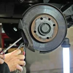 Person measuring brake pad thickness beside a car wheel removed in a home garage